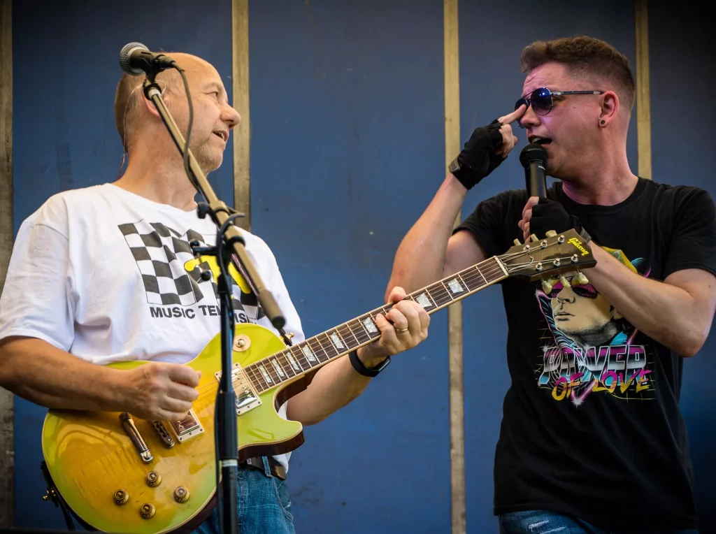 Two members of the 80s tribute band Regener8 perform on stage. The guitarist, wearing a white checkered t-shirt, plays a lemon burst Les Paul guitar while smiling at the lead singer, who is holding a microphone and adjusting his sunglasses mid-performance.