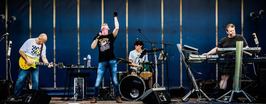 The full Regener8 band performs live outdoors against a plain blue backdrop. The guitarist plays a bright yellow Les Paul, the singer stands centre stage with a fist raised, the drummer keeps rhythm behind a green kit, and the keyboardist plays on a dual-tiered setup while wearing a kilt.