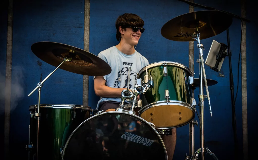 A young drummer from the 80s tribute band Regener8 smiles while performing on a green drum kit. He wears dark sunglasses and a light-coloured T-shirt with a music-themed graphic, seated in front of a blue backdrop at an outdoor event.