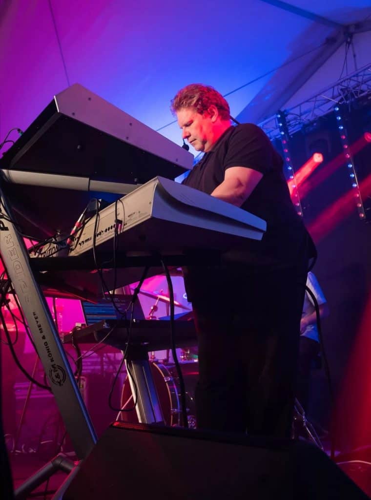 A keyboardist from the 80s tribute band Regener8 performs on stage with a dual keyboard rig. He is focused, wearing a black shirt and headset mic, lit by pink, blue, and red stage lights beneath a marquee tent.