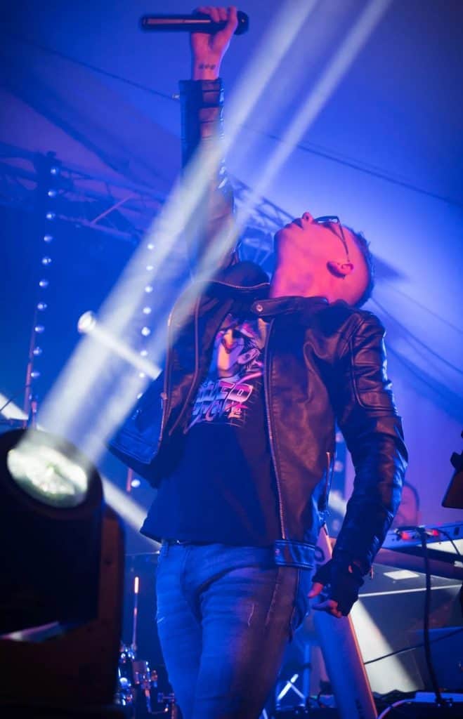 Regener8’s lead vocalist holds a microphone high in the air while looking upward, caught in a dramatic moment on stage. Dressed in a black leather jacket and retro graphic tee, he stands illuminated by powerful white beams of stage lighting cutting through the blue haze.