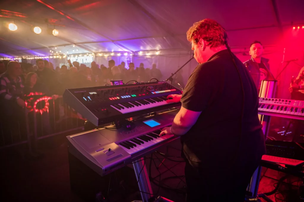 A keyboard player from the 80s tribute band Regener8 performs on stage under warm pink and purple lighting. He is playing a dual keyboard setup, facing a large crowd of engaged audience members inside a marquee filled with string lights and vibrant atmosphere.