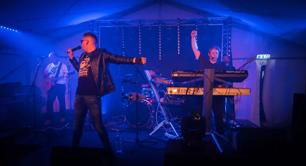 A live performance photo of the 80s tribute band Regener8 on stage under blue and purple lighting. The lead singer stands front and centre in a leather jacket and sunglasses, extending his arms dramatically with a microphone in hand. A keyboardist raises a fist while playing, with the drummer, guitarist, and bassist performing in the background.