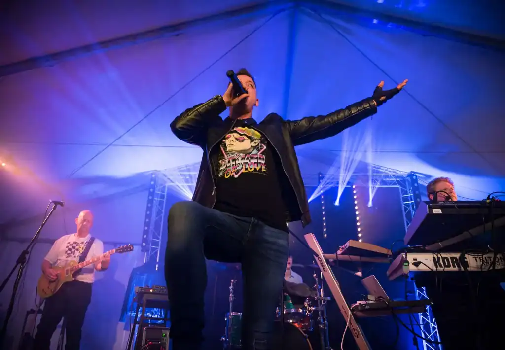 A powerful low-angle shot of Regener8's lead singer performing on stage, one knee raised, with his arm outstretched and microphone in hand. Stage lights beam behind him as the guitarist, drummer, and keyboardist play under a white marquee tent with dramatic lighting.