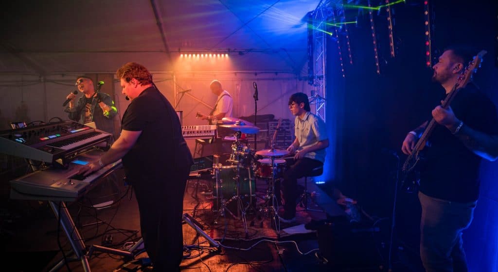 The full Regener8 band performs live inside a marquee tent. The keyboardist plays a dual setup in the foreground, while the drummer, bassist, guitarist, and lead singer are positioned across the stage. Colourful stage lighting fills the space with warm and cool tones.