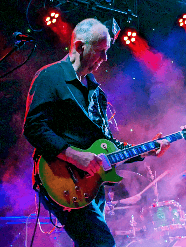 A guitarist from the 80s tribute band Regener8 performs live on stage, focused as he plays a sunburst green Les Paul electric guitar. He’s surrounded by vibrant red and purple stage lights with fog effects, creating a dramatic rock atmosphere.