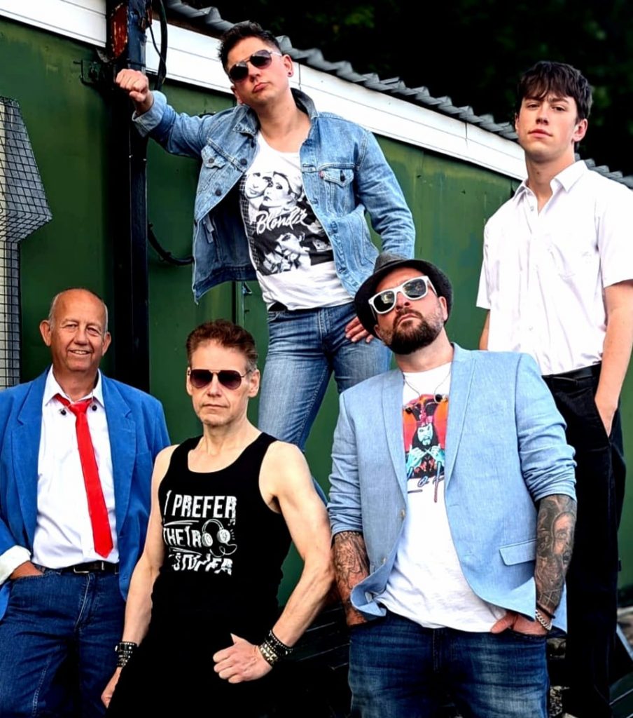 A group photo of five male band members posing outside in front of a green and white structure. Each person is dressed in a distinctive 80s-inspired outfit, featuring denim, sunglasses, printed T-shirts, and vibrant accessories. The central figure stands on a step, leaning against a metal pole.