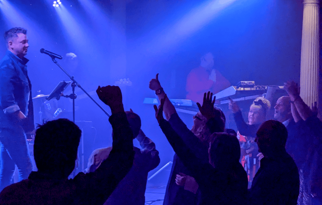 Regener8's lead singer stands at the microphone on a misty, blue-lit stage as the crowd in the foreground raises their hands, cheering and pointing toward the band. The keyboardist can be seen performing in the background, dressed in red.