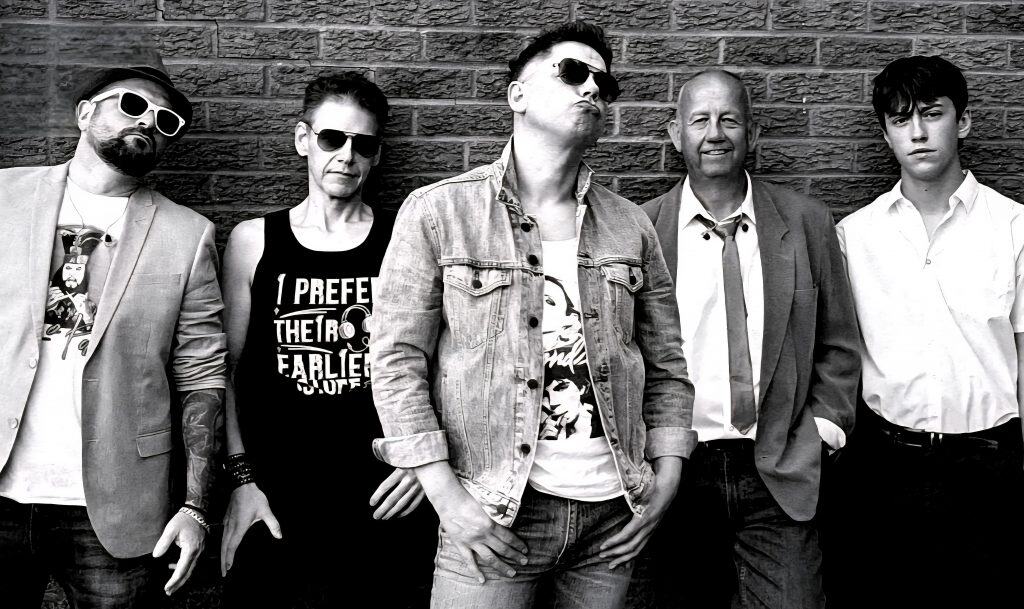 Regener8 band members posing in front of a brick wall in black and white. Each member showcases unique 80s-inspired style with sunglasses, graphic tees, and denim jackets.